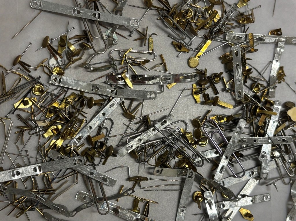A collection of various metal fasteners and pins, including safety pins, thumbtacks, and paper clips, scattered on a light surface.