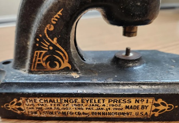 Close-up of a vintage eyelet press labeled 'THE CHALLENGE EYELET PRESS N°1' with patent dates and manufacturer details.
